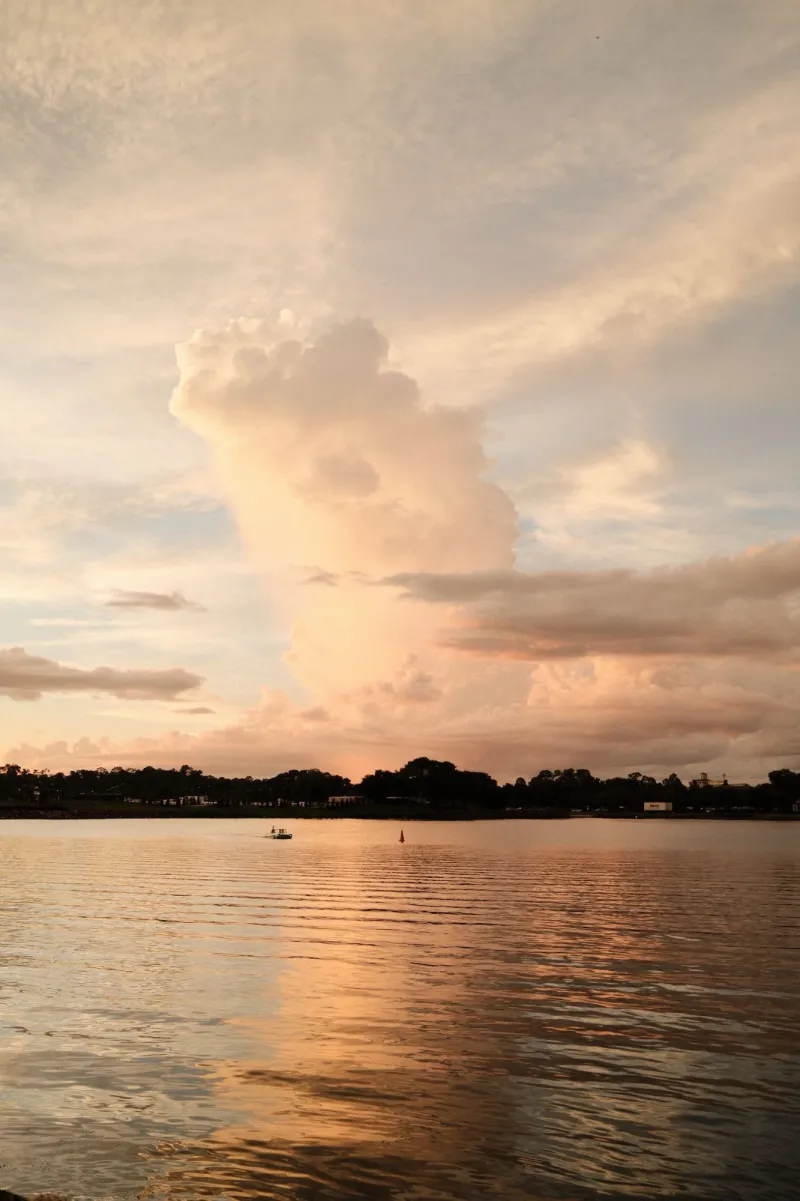 A tall sunset cloud glowing peach over a calm lake, a small boat crossing below a tree line.