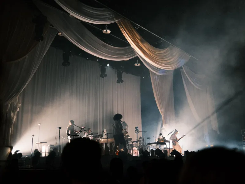 A band performing on a haze-filled stage under swags of draped cream fabric, seen over the crowd's silhouettes.