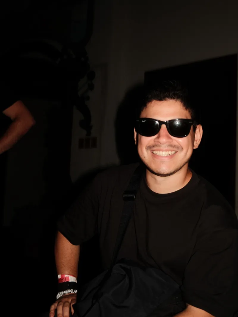 Jason smiling in sunglasses and a black T-shirt, lit by flash against a dark room.