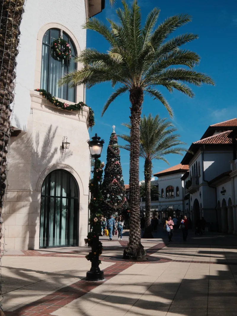 Palm trees and a decorated holiday tree in a sunny plaza of white stucco buildings and red tile roofs.