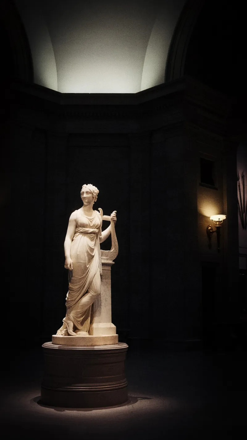 A marble statue of a woman holding a lyre, softly spotlit in a dark museum gallery.