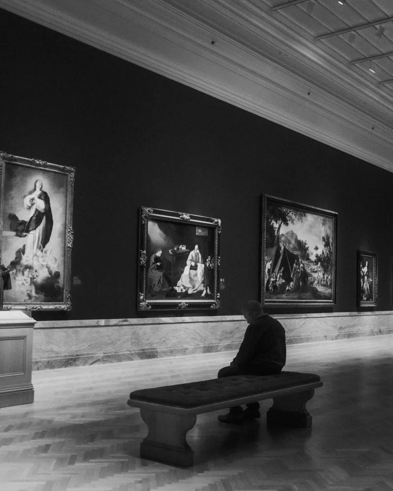 Black-and-white museum gallery, a lone visitor on a bench facing large framed paintings.