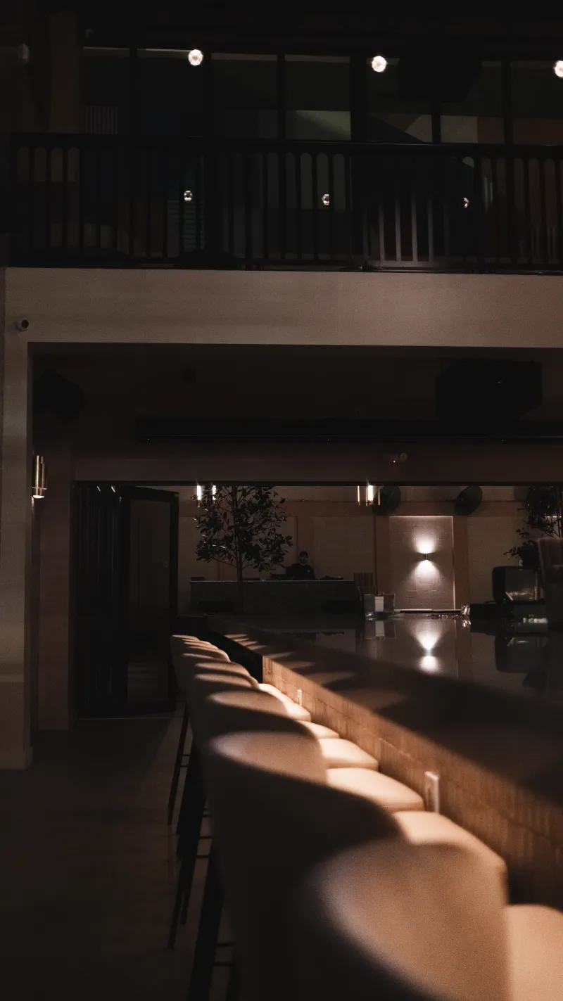 A dim, empty bar at night, warm light catching a long row of rounded stools beneath a balcony.