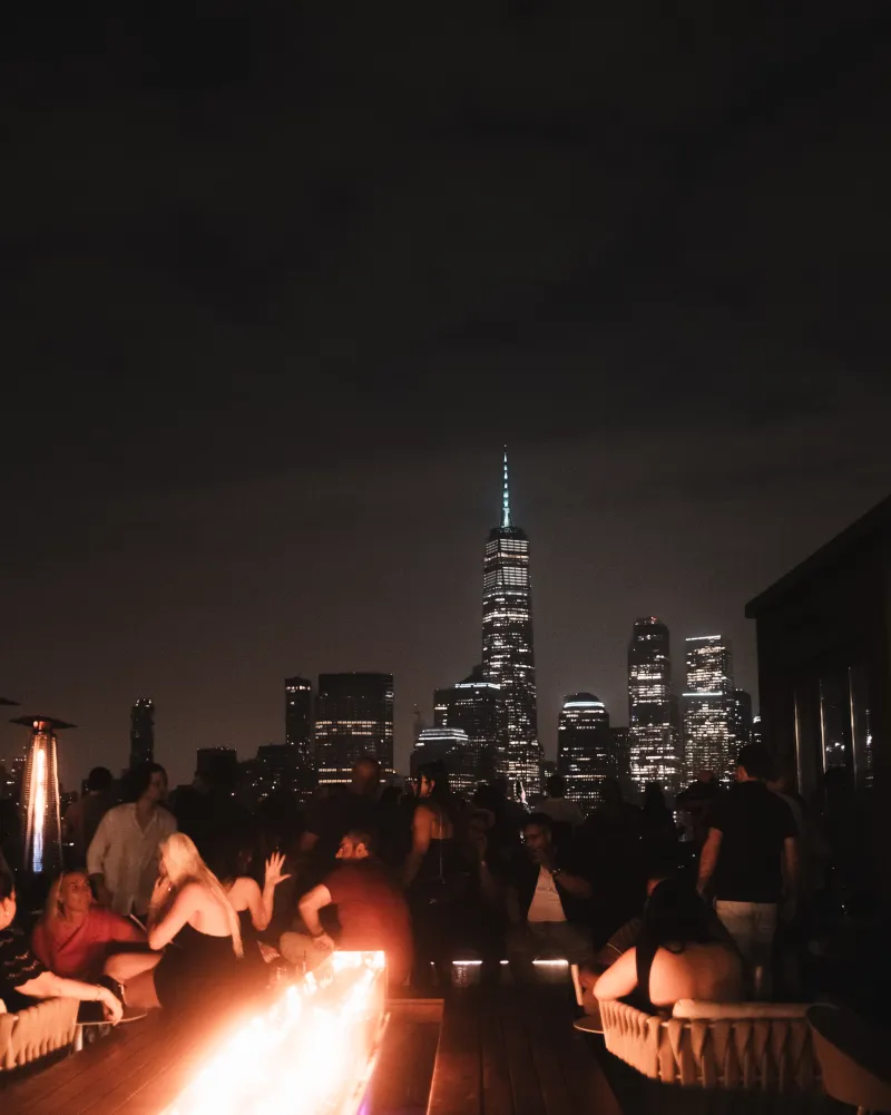 People around a glowing rooftop fire table at night, a lit city skyline and tall spire beyond.