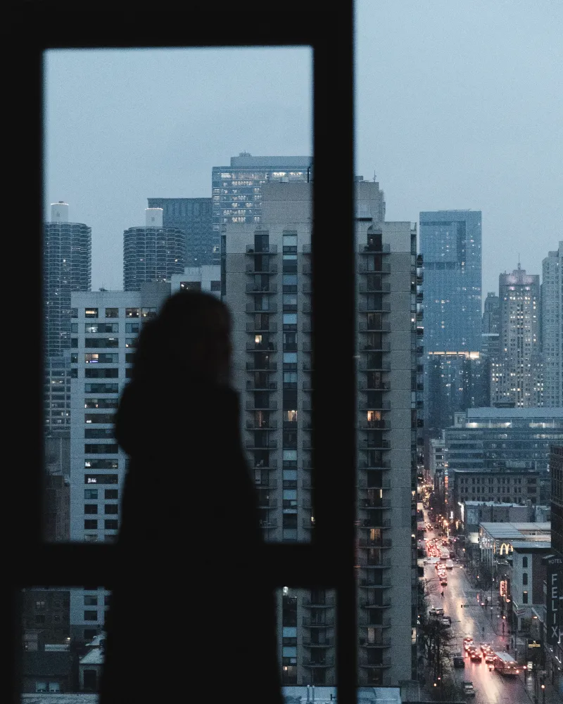 A figure silhouetted at a tall window, looking over city towers and a street of red tail lights at dusk.