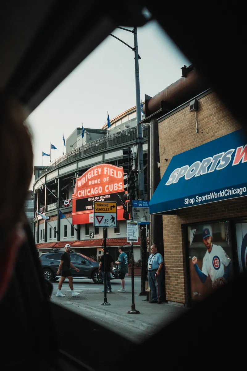 A ballpark's red marquee seen through a car window, people crossing the street below.