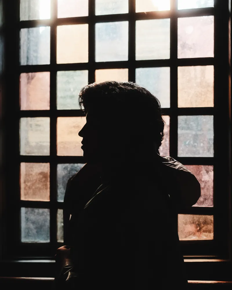 A person in profile, silhouetted against a tall leaded-glass window glowing with soft light.