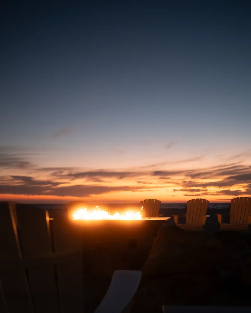 Sunset over the sea, a fire table glowing beside a row of empty Adirondack chairs.